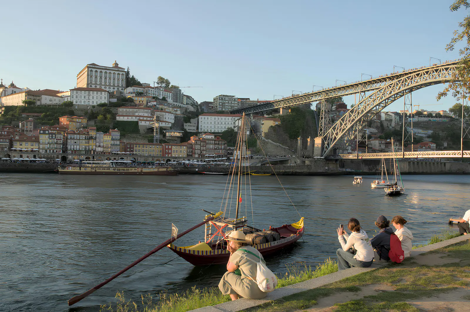People resting by the Douro with Dom Luís I Bridge and a rabelo boat nearby.